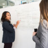 two women conversing in front of a whiteboard