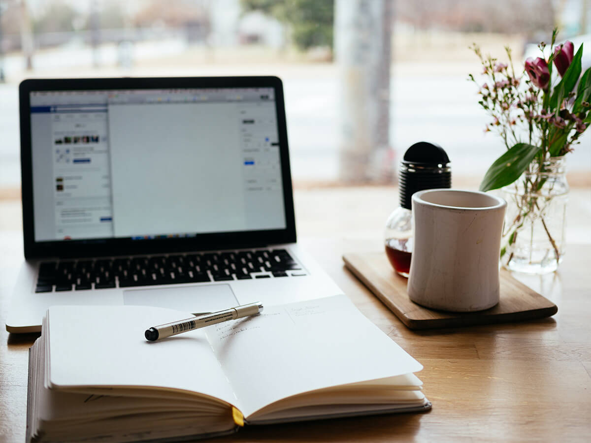 laptop, notebook and pen, and a coffee mug on a table overlooking a window