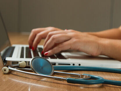 woman typing on a laptop with a stethoscope on desk next to the laptop